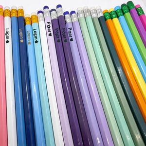 May include: A collection of colorful pencils in various shades, including pink, blue, purple, green, and yellow. Each pencil has a white barrel with text and a heart symbol. The pencils have erasers and gold-colored metal bands.