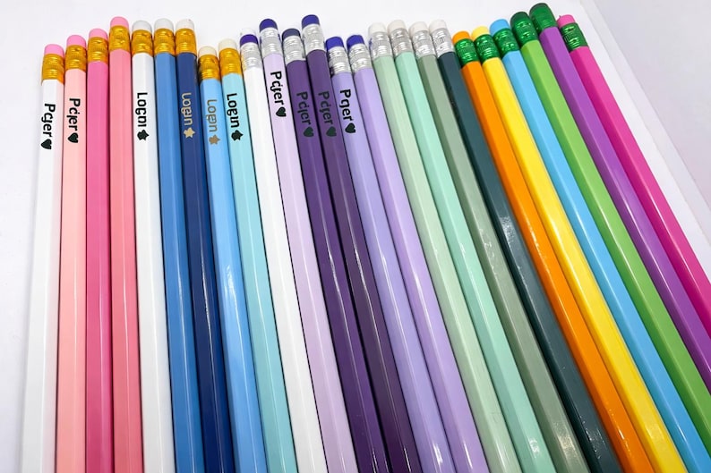 May include: A collection of colorful pencils in various shades, including pink, blue, purple, green, and yellow. Each pencil has a white barrel with text and a heart symbol. The pencils have erasers and gold-colored metal bands.