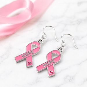 May include: A pair of silver-toned drop earrings featuring a pink enamel awareness ribbon design. Each ribbon charm is engraved with the word HOPE in capital letters and includes a small pink heart detail at the bottom of the ribbon loop.
