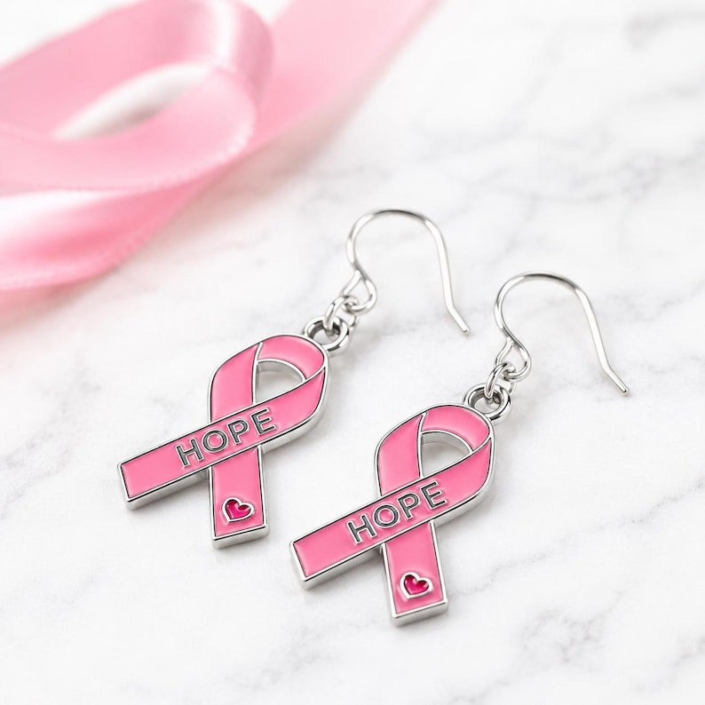 May include: A pair of silver-toned drop earrings featuring a pink enamel awareness ribbon design. Each ribbon charm is engraved with the word HOPE in capital letters and includes a small pink heart detail at the bottom of the ribbon loop.