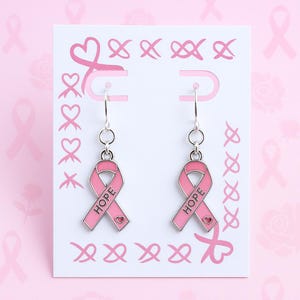 May include: A pair of silver-toned dangle earrings featuring a pink enamel ribbon charm with the word HOPE printed in black and a small heart detail at the bottom. The earrings are displayed on a white card decorated with pink ribbon patterns.