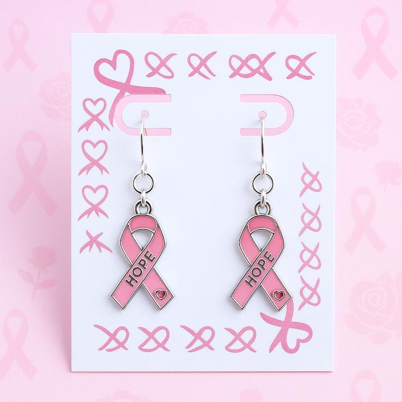 May include: A pair of silver-toned dangle earrings featuring a pink enamel ribbon charm with the word HOPE printed in black and a small heart detail at the bottom. The earrings are displayed on a white card decorated with pink ribbon patterns.