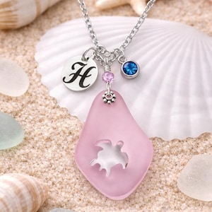 Könnte beinhalten: Eine silberne Halskette mit einem rosa Seeglas-Anhänger mit einem Schildkröten-Ausschnitt. Die Halskette hat einen Anhänger mit dem Buchstaben "H", einen blauen Edelstein und rosa Perlen. Der Hintergrund zeigt Muscheln und Sand.