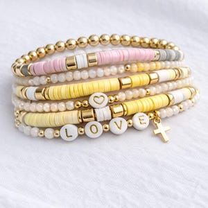 May include: Stack of beaded bracelets in pastel colors, including pink, yellow, and white. Some bracelets feature gold accents, heart-shaped beads, and letter beads spelling "LOVE". A gold cross charm hangs from one bracelet.