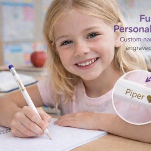 May include: A white pencil with a purple eraser and the name "Piper" and a gold heart engraved on it. The pencil is being used to write on a spiral notebook. The image also includes the text "Fun Personalization! Custom name & heart engraved."