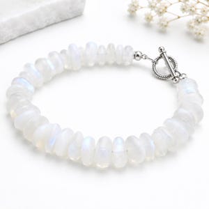 May include: A bracelet featuring white moonstone beads and a silver toggle clasp. The irregular, polished moonstone beads have a translucent, milky appearance with iridescent flashes. The clasp is a simple, elegant design. A piece of jewellery.