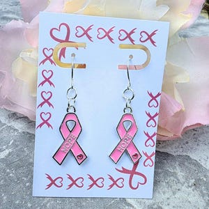 May include: A pair of silver-toned hook earrings featuring pink enamel awareness ribbons. Each ribbon charm is embossed with the word HOPE and a small heart detail. The earrings are displayed on a white card decorated with a pink ribbon pattern.