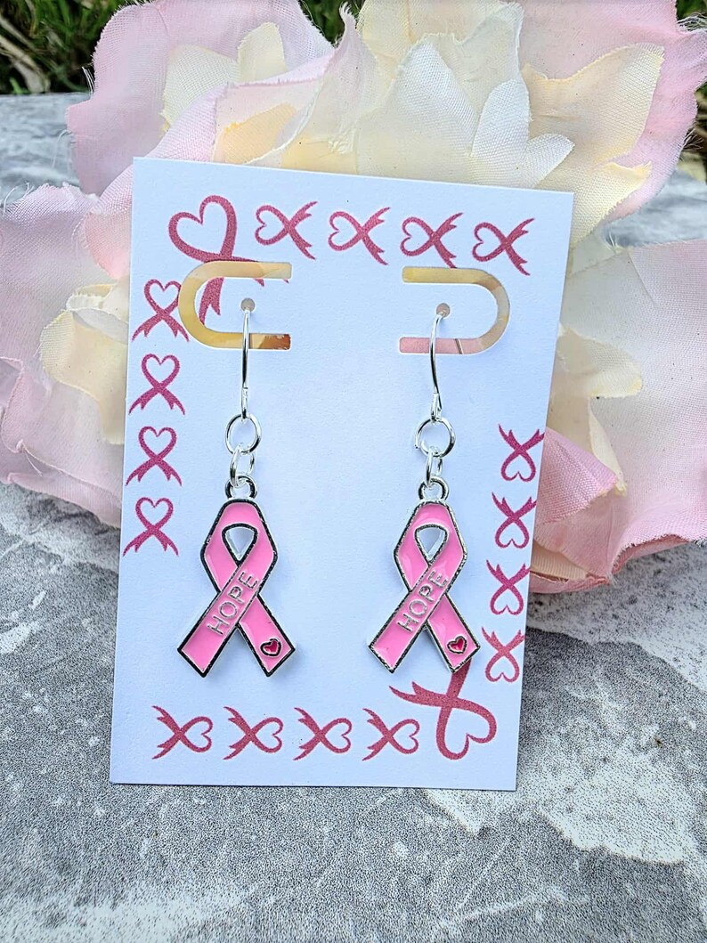 May include: A pair of silver-toned hook earrings featuring pink enamel awareness ribbons. Each ribbon charm is embossed with the word HOPE and a small heart detail. The earrings are displayed on a white card decorated with a pink ribbon pattern.