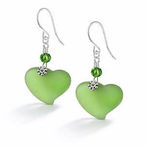 Pode incluir: Um par de brincos em forma de coração. Cada brinco apresenta um pingente de coração verde claro fosco suspenso de um gancho de prata. Pequenas contas verdes e detalhes florais prateados adicionam detalhes. Fundo branco.