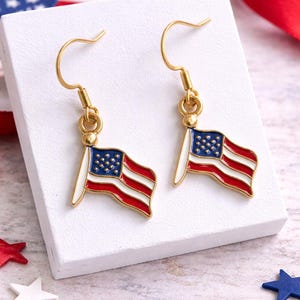 Puede incluir: Pendientes dorados con banderas americanas en miniatura. Cada bandera tiene un campo azul con estrellas blancas, rayas rojas y blancas y un mástil blanco. Los pendientes se exhiben sobre una superficie blanca con decoraciones en forma de estrella.