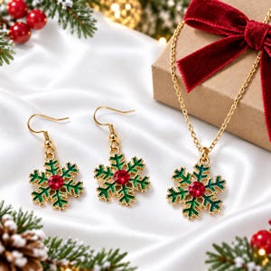 May include: A gold-tone jewelry set consisting of a necklace and two drop earrings. Each piece features a snowflake-shaped pendant with green enamel detailing and a central round red crystal. The necklace includes a delicate gold-tone chain.