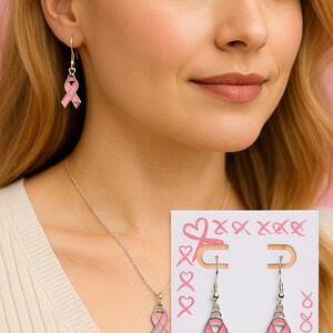 May include: A silver-toned necklace and earring set featuring pink ribbon pendants. Each ribbon charm is enameled in light pink with the word HOPE and a small heart detail. The earrings are displayed on a white card decorated with pink ribbon and heart illustrations.