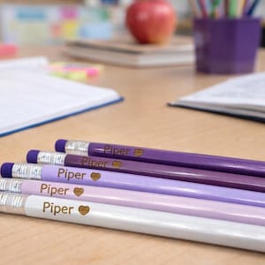 May include: A collection of personalized pencils in shades of purple and white, each labeled "Piper" with a heart symbol. The pencils are arranged on a wooden desk, with school supplies in the background.