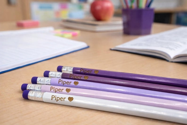 May include: A collection of personalized pencils in shades of purple and white, each labeled "Piper" with a heart symbol. The pencils are arranged on a wooden desk, with school supplies in the background.