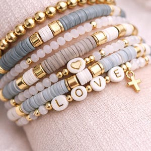 May include: A stack of beaded bracelets in shades of gray, white, and gold. Some bracelets feature the word "LOVE" spelled out with white letter beads. One bracelet has a gold cross charm. The bracelets are stacked on a light beige surface.