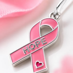 May include: A silver-toned metal awareness ribbon pendant featuring a smooth pink enamel inlay. The word HOPE is embossed in silver capital letters across the center of the ribbon, and a small pink heart shape is positioned at the bottom tail.