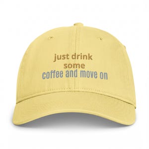 Pode incluir: Um boné de beisebol amarelo-pálido com texto bordado no painel frontal. As palavras "just drink" e "some" são costuradas com linha castanho-escura, enquanto "coffee and move on" são costuradas com linha azul-clara.