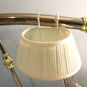 May include: Cream-colored pleated lampshade hanging from a brass-colored metal frame. The lampshade has a rounded, tapered shape and is suspended by wire hooks. The metal frame has decorative gold accents.
