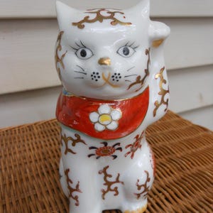 May include: A white ceramic Maneki Neko cat figurine with gold accents. The cat is depicted with one paw raised, wearing a red collar with a white flower. The figurine is decorated with gold floral patterns and has black whiskers.