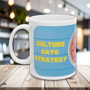 Op de afbeelding: Een witte keramische mok met een lichtblauwe achtergrond met een horizontale houtnerfstructuur. De tekst "CULTURE EATS STRATEGY" is gedrukt in dikke gele hoofdletters. Een deel van een roze geglazuurde donut met kleurrijke hagelslag is zichtbaar.