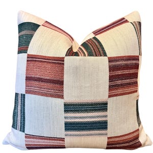 Op de afbeelding: Vierkant decoratief kussen met een patchwork ontwerp. Het kussen heeft een crèmekleurige achtergrond met vierkanten van rood, groen en crèmekleurig gestreepte stof. Het kussen heeft een vierkante vorm.