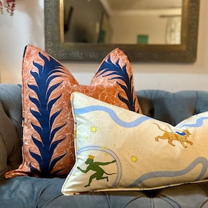 Może przedstawiać: Trzy dekoracyjne poduszki na szarej kanapie. Poduszki są wykonane z tkaniny o r&oacute;żnych wzorach. Jedna poduszka ma wz&oacute;r w kolorze niebieskim i pomarańczowym z liściastym wzorem. Inna poduszka ma kremowy kolor z niebieską rzeką, gwiazdami i zieloną małpą z koroną. Trzecia poduszka ma kremowy kolor z niebieską rzeką, gwiazdami i brązowym gepardem.