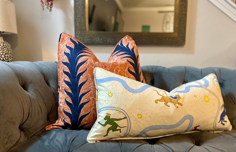 Może przedstawiać: Trzy dekoracyjne poduszki na szarej kanapie. Poduszki są wykonane z tkaniny o r&oacute;żnych wzorach. Jedna poduszka ma wz&oacute;r w kolorze niebieskim i pomarańczowym z liściastym wzorem. Inna poduszka ma kremowy kolor z niebieską rzeką, gwiazdami i zieloną małpą z koroną. Trzecia poduszka ma kremowy kolor z niebieską rzeką, gwiazdami i brązowym gepardem.