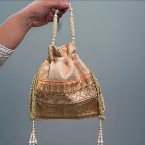 May include: A peach-coloured drawstring pouch bag featuring intricate gold embroidery and sequin detailing across the lower section. The bag includes a handle made of white faux pearls and two long gold braided drawstrings with pearl-beaded tassels.