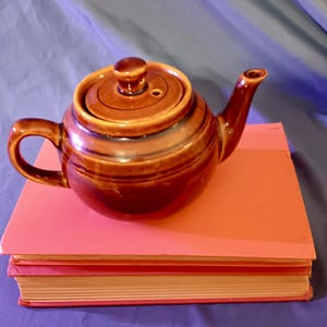 May include: A glossy brown ceramic teapot with a matching lid, featuring a curved handle and a tapered spout. The teapot has a rounded body with horizontal ridged detailing around the center and sits atop a stack of two orange books.