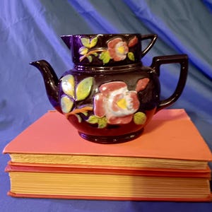 May include: A dark brown ceramic teapot featuring a raised, hand-painted floral design with pink and white petals and green leaves. The teapot has a curved spout, a sturdy handle, and a matching lid with a small handle, resting on two books.