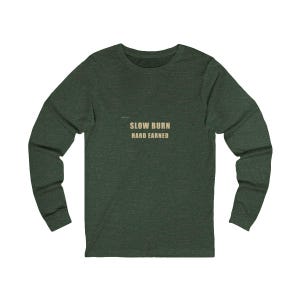 Op de afbeelding: Een donker bosgroen shirt met lange mouwen en de tekst SLOW BURN HARD EARNED in een lichtbeige kleur op het midden van de borst. Het kledingstuk heeft een ronde hals en lange mouwen met geribbelde boorden, voorzien van een gemêleerde textuur.