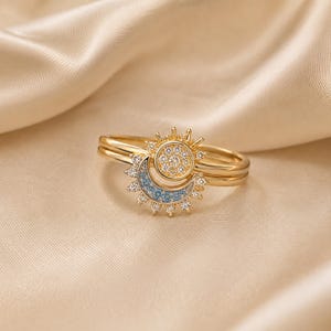 Puede incluir: Un anillo de tono dorado que presenta un motivo de sol circular central incrustado con piedras claras, junto con una forma de media luna acentuada con piedras azul claro. El diseño de sol y luna está rodeado por pequeñas puntas de piedras claras.