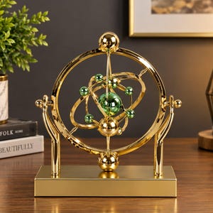May include: A kinetic desk sculpture featuring a gold-coloured metal frame and rotating rings. At the centre, a large green sphere is surrounded by smaller green spheres, all suspended within the gold structure on a rectangular gold base.