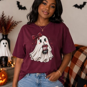 May include: A maroon short-sleeve t-shirt featuring a graphic of a white ghost wearing a dark red bow and holding a can of Dr Pepper Zero Sugar. The ghost has blushing cheeks and heart-shaped eyes, surrounded by small red hearts and sparkling star accents.