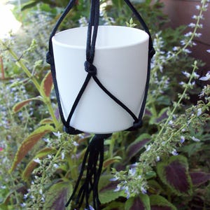 May include: A white cylindrical plant pot suspended in a black macrame hanger. The hanger is constructed from black cord with knotted details that secure the pot in place, ending in a tassel of loose cord strands hanging below the base.