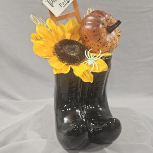 May include: A black ceramic boot-shaped vase filled with a large yellow sunflower, a speckled brown pumpkin, and a small light blue spider. A miniature wooden ladder stands behind the flowers, featuring a white sign labeled Pumpkin Patch.