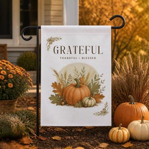 Op de afbeelding: Een rechthoekige witte tuin vlag hangend aan een zwarte metalen standaard. De vlag bevat de tekst "GRATEFUL" in grote donkere letters, met "THANKFUL • BLESSED" eronder. Een illustratie toont een grote oranje pompoen, een kleine witte pompoen en tarwe.