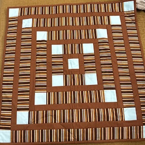 May include: A square quilt featuring a geometric log cabin pattern. The design consists of alternating strips of brown, tan, and white striped fabric, solid brown fabric borders, and light blue square accents arranged in a concentric layout.