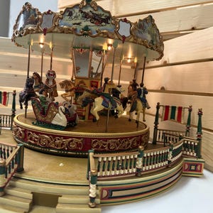 May include: A decorative musical carousel featuring a circular platform with miniature horses and a swan-shaped sleigh. The structure includes gold-toned ornate detailing, a central pillar with mirrors, and a tiered base with green and red railings.