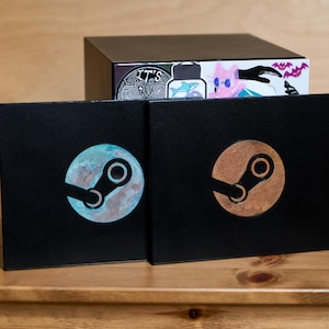 May include: Two black square cases featuring the Steam logo. The left case has a blue and white marbled circular logo, while the right case has a copper-toned circular logo. Behind them sits a black box decorated with various colourful stickers.