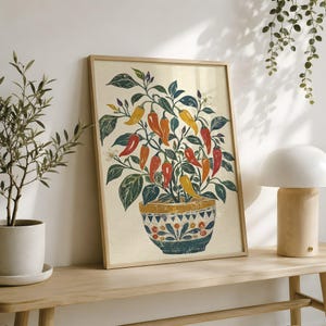 May include: A framed wall art print featuring a potted chili pepper plant with vibrant red, orange, and yellow peppers and dark green leaves. The plant sits in a decorative bowl with a blue, white, and orange geometric pattern on a cream background.