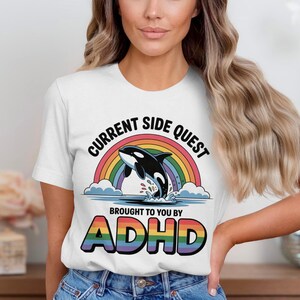 Op de afbeelding: Een wit t-shirt met korte mouwen en een afbeelding van een zwart-witte orka die over blauw water springt voor een kleurrijke regenboog. De tekst luidt CURRENT SIDE QUEST BROUGHT TO YOU BY ADHD in dikke zwarte en regenboogkleurige letters.