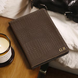 May include: A rectangular dark brown notebook with a textured crocodile skin pattern. The cover features the gold embossed initials G.R in the bottom right corner. The notebook is closed and has a slim profile with visible stitching along the edges.