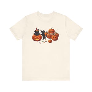 May include: A cream-colored t-shirt featuring a graphic of two black cats, one wearing a witch hat inside a jack-o'-lantern, and another playing with a ghost. The design includes several orange pumpkins, a candle, candy, and the text SPOOKY TRICK-OR-TREAT BOO!