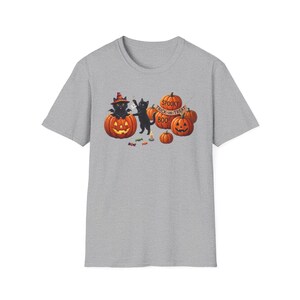 Peut inclure: Un t-shirt gris chiné avec une illustration de deux chats noirs, dont l'un porte un chapeau de sorcière, assis parmi des citrouilles orange. Une bannière sur les citrouilles indique "SPOOKY TRICK-OR-TREAT BOO!" avec des bonbons éparpillés.