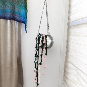 May include: A spherical disco ball planter hanging from a silver metal chain. The planter contains crocheted trailing vines in dark green, white, and light pink, featuring small round pom-pom accents that dangle downward from the reflective surface.