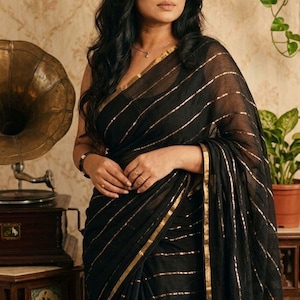 May include: A black saree featuring horizontal gold metallic stripes and a solid gold border. The fabric appears lightweight and semi-sheer, draped elegantly over the shoulder. The garment is styled with a simple black sleeveless blouse underneath.
