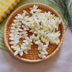May include: A circular woven rattan basket containing a coiled garland of fresh white jasmine flowers. The delicate blossoms are strung together with thin white thread, displaying small pale yellow centers and soft, rounded white petals.