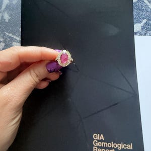 May include: A gold ring featuring an oval-cut pink gemstone surrounded by a halo of small white diamonds, held against a black GIA Gemological Report document with the GIA logo and text printed in white and gold at the top and bottom.