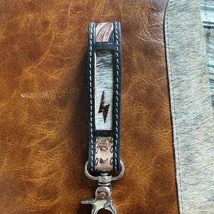 May include: A leather keychain featuring a central panel of white and black cowhide with a dark brown lightning bolt graphic. The strap is bordered by black leather with white stitching and includes a silver-toned metal swivel lobster clasp.